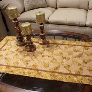 Könnte beinhalten: Ein goldener Tischläufer mit einem geometrischen Muster aus Dreiecken. Der Läufer liegt auf einem Couchtisch mit Glasplatte und drei goldenen Kerzenständern mit Kerzen.