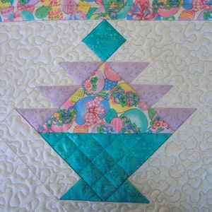 以下が含まれることがあります： ターコイズのバスケットとラベンダーと模様のトップが特徴の、キルト加工されたイースターバスケットのデザイン。バスケットは、イースターエッグとリボンの装飾的なボーダーが付いた白い背景に配置されています。キルトはミシン縫いです。