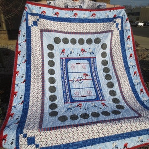 May include: A blue and red hockey themed quilt with a hockey rink design in the center. The quilt has a white background with blue and red borders. The text "SLAP SHOT POWER PLAY FACE OFF OVERTIME" is printed on the quilt.