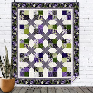 May include: A purple and green quilt with a floral pattern. The quilt is made up of squares and has a white background. The quilt is hanging on a white brick wall.