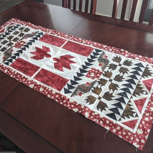 Puede incluir: Un camino de mesa acolchado rojo, blanco y negro con un patrón geométrico. El camino de mesa presenta un diseño central de una hoja de arce estilizada, rodeada de formas geométricas más pequeñas.
