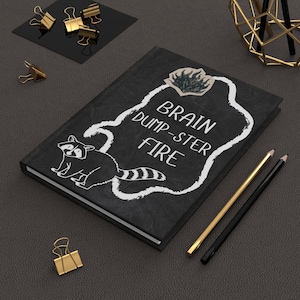 May include: A black hardcover notebook featuring a white outline of a raccoon and a flame graphic. The text reads BRAIN DUMP-STER FIRE in white lettering. The notebook is accompanied by two pencils and several gold-colored metal binder clips.