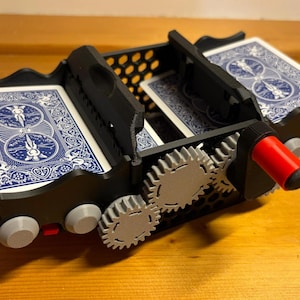 Op de afbeelding: Een zwarte plastic kaartenschudder met een mechanisch tandwielsysteem met grijze tandwielen en een rode hendel. Het apparaat bevat twee stapels speelkaarten met blauw-witte patronen op de achterkant en heeft een honingraatstructuur aan de zijkanten.