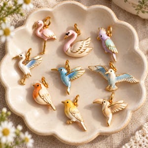 May include: A collection of nine gold-toned metal charms featuring colourful enamel bird designs, including a pink flamingo, a white swan, a purple parrot, a white crane, a blue hummingbird, a white seagull, a robin, a yellow bird, and a toucan.