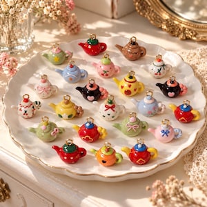 May include: A collection of miniature teapot charms in various colors and patterns, including red, blue, yellow, pink, green, and black. Each small ceramic-style teapot features a gold-tone metal loop at the top for hanging as a jewelry pendant.