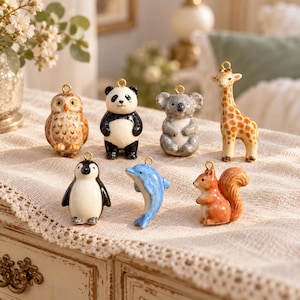 May include: A collection of seven small ceramic animal charms featuring an owl, panda, koala, giraffe, penguin, dolphin, and squirrel. Each figurine has a gold-toned metal loop at the top for hanging, showcasing detailed painted features.
