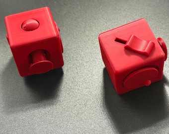 May include: Two bright red fidget cubes featuring various tactile elements on each side. One cube displays a circular button and a protruding peg, while the other shows a sliding switch and a circular dial. Both cubes have a matte finish.