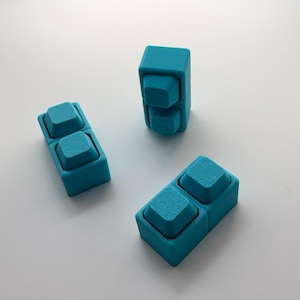 May include: Three bright teal rectangular blocks, each featuring two square, slightly raised buttons on the top surface. The blocks have a matte finish and a structured, geometric design, arranged in a scattered formation on a plain surface.