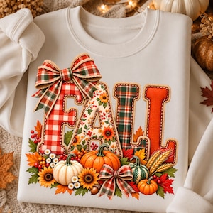 May include: A cream-colored sweatshirt featuring the word FALL in large, textured letters. The F is red and white checkered with a plaid bow, the A has a floral pattern, and the Ls are plaid and orange. Pumpkins, sunflowers, and leaves surround the text.