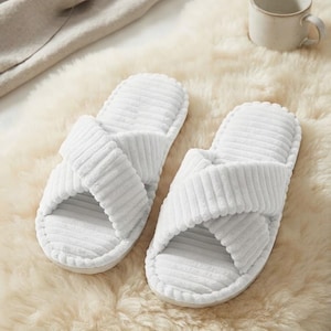 May include: A pair of white open-toe slippers featuring a textured corduroy-style fabric. The design consists of two wide, crisscrossing straps over the top of the foot and a matching cushioned footbed with a rounded toe and heel shape.