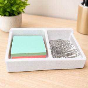 May include: A rectangular white desk organizer with a speckled texture featuring two compartments. One side holds a stack of square light green and pink sticky notes, while the other side contains a collection of silver metal paper clips.