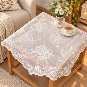 Peut inclure: Une nappe carrée en dentelle blanche ornée d'un motif floral complexe avec des bords festonnés. Le tissu présente une texture tissée semi-transparente. La nappe est posée sur une table en bois, offrant un revêtement décoratif pour la surface.