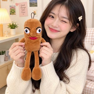 May include: A brown plush toy shaped like a sausage with a cartoon face, featuring large white eyes with black pupils, a prominent nose, and bright red lips. The soft toy has thin arms and long legs with rounded feet and a small black hanging loop.