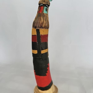 May include: A hand-carved wooden figurine featuring a tall, slender body painted with horizontal bands of red, yellow, and black. The figure has a textured, wood-grain head with turquoise-coloured feather accents and a natural wood base.