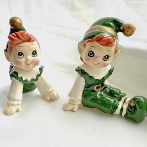 Könnte beinhalten: Zwei Keramik-Elfenfiguren mit glänzender Oberfläche. Beide Figuren haben orangefarbenes Haar, spitze grüne Mützen mit goldenen Spitzen, weiße Kragen und grüne Kleidung. Eine Figur krabbelt, während die andere mit ausgestreckten Beinen sitzt.