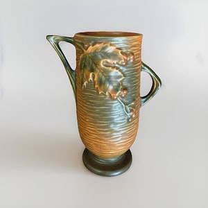 May include: A ceramic pitcher featuring a textured, horizontal ribbed surface in shades of brown and green. The body is adorned with a raised relief of a large leaf and small berries. It has a pedestal base and two asymmetrical handles.