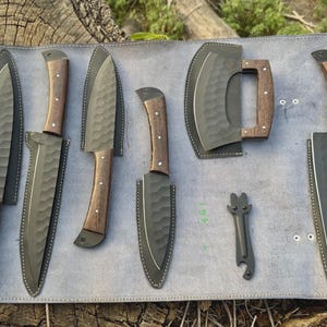 May include: A set of five kitchen knives and a small tool with dark, hammered-finish blades and brown wooden handles, each stored in a black leather sheath, arranged on a light gray leather roll-up mat with visible stitching and metal rivets.