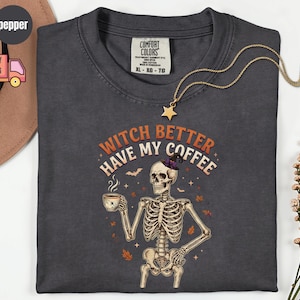 Può includere: Una t-shirt grigio antracite con la grafica di uno scheletro che indossa un cappello da strega e tiene in mano una tazza di caffè fumante. Il testo "WITCH BETTER HAVE MY COFFEE" è scritto in arancione e bianco, circondato da pipistrelli e foglie.