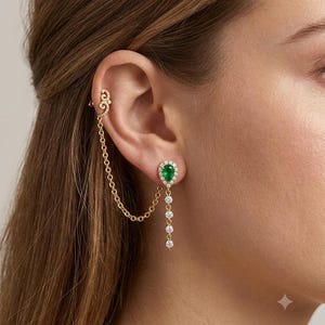 Könnte beinhalten: Ein goldener Ohrring und ein Ear Cuff, die durch eine feine Goldkette verbunden sind. Der Ear Cuff hat ein geschwungenes Design, während der Ohrstecker einen birnenförmigen grünen Edelstein mit weißem Steinbesatz und einen hängenden Kettenanhänger zeigt.