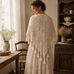 May include: A cream-colored triangular crochet shawl featuring an intricate openwork pattern of floral motifs and geometric shapes. The edges of the shawl are adorned with small, dangling tassels that hang from the bottom and sides of the garment.