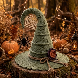 Könnte beinhalten: Ein spitzer Häkelhut in Salbeigrün mit breiter, gewellter Krempe. Der Hut ist mit einem hängenden weißen Häkel-Spinnennetz samt schwarzer Spinne, einer dicken, zu einer Schleife gebundenen Juteschnur und einer dunkelviolett-orangen Häkelblume verziert.