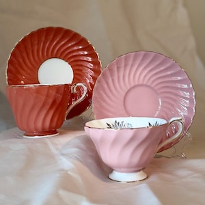 May include: Two tea cups and matching saucers with a swirl pattern. One set is a deep coral orange with a white centre on the saucer, and the other is a soft pastel pink. Both cups feature gold-toned rims and handles with a white base.