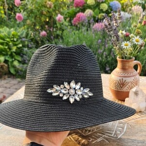Op de afbeelding: Een zwarte strooien fedora-hoed met een decoratieve cluster van heldere, gefacetteerde kristallen in een marquise- en rond patroon aan de voorzijde. De hoed heeft een gestructureerde kroon en een brede, licht neerwaartse rand met textuur.