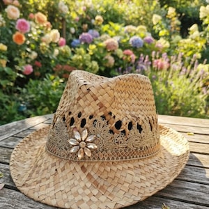 May include: A tan woven straw fedora hat featuring a wide brim and a pinched crown. The hat is decorated with a band of intricate beige lace trim and a central flower-shaped embellishment made of clear, faceted crystals set in a metallic base.