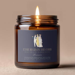 May include: A lit, amber-colored glass jar candle with a navy blue label featuring an illustration of three figures. The label reads "LYME REGIS ON THE COBB" and "Persuasion". The candle is scented and weighs 8.7 oz.