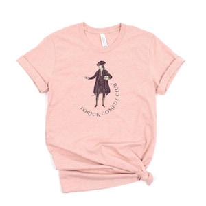 May include: A light pink t-shirt featuring a vintage illustration of a man in a hat and coat, with the text "Yorick Comedy Club" in a curved design. The shirt has rolled-up sleeves and a classic crew neck.