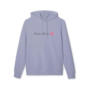 Peut inclure: Un sweat-shirt à capuche violet clair avec une poche kangourou sur le devant et des manches longues. La poitrine porte l'inscription "Made with love" dans une police cursive gris foncé, accompagnée d'une petite icône de cœur rose uni.