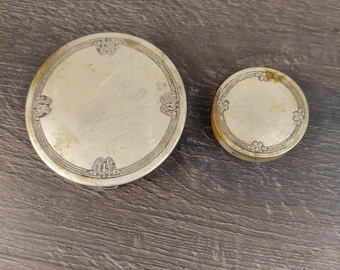 May include: Two round, metallic compact cases with a gold-toned finish. The larger case features a decorative border with four symmetrical engraved motifs, while the smaller case has a similar circular design with a matching engraved pattern.