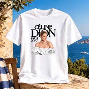 Pode incluir: Uma t-shirt branca de manga curta com uma impressão gráfica de Céline Dion. O texto CÉLINE DION PARIS 2027 aparece acima da imagem, e L'ORÉAL PARIS OFFICIAL BEAUTY PARTNER está impresso abaixo do retrato da pessoa com roupa branca.