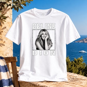 May include: A white short-sleeved t-shirt featuring a black and white photographic print of Celine Dion centered on the chest. The text CELINE is printed above the portrait and DION is printed below, both in a distressed, block-letter font.