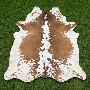 May include: A cowhide rug featuring a natural pattern of brown and white patches. The hide has an irregular, organic shape with a central white stripe running vertically, surrounded by large brown sections and smaller white spots throughout.