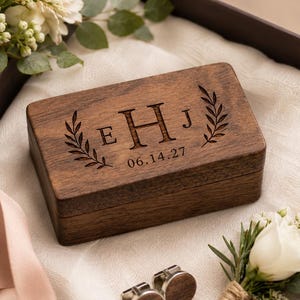 May include: A rectangular dark wood box with a hinged lid, engraved with the letters E, H, and J, a laurel leaf design, and the date 06.14.27. Two circular wood-inlay cufflinks are positioned nearby, accompanied by white floral accents.