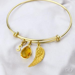 May include: A gold bangle bracelet with three charms: a letter 'S', a teardrop-shaped amber gemstone, and a gold angel wing.