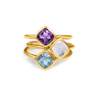 Pode incluir: Três anéis de ouro com pedras preciosas de forma quadrada. O anel superior tem uma ametista roxa, o anel do meio tem uma pedra da lua branca e o anel inferior tem uma água-marinha azul claro.