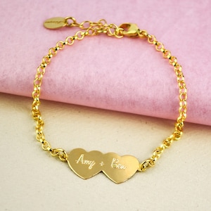 May include: A gold-colored bracelet with a chain design. The bracelet features two heart-shaped pendants with the names "Amy + Ben" engraved on them. The bracelet is displayed on a pink surface.