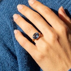 May include: A gold ring with a large, oval-shaped blue gemstone. The ring is set on a hand, with a blue sweater in the background. The ring's setting appears to be simple, allowing the gemstone to be the focal point.