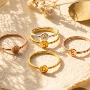 May include: Four thin metal rings featuring small circular charms, each engraved with a single letter. One ring is silver-toned with the letter A, while the other three are gold-toned and feature the letters B, C, D, and E respectively.