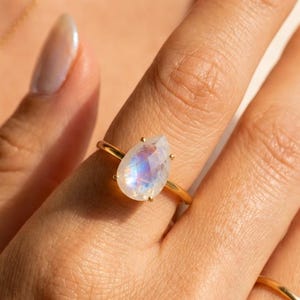 Moonstone teardrop ring worn on hand in warm sunlight, featuring a pear-shaped rainbow moonstone with blue flash in a prong setting on a gold band. A June birthstone ring, gemstone statement ring, gift for her, birthday gift, anniversary gift, or meaningful everyday jewelry piece.