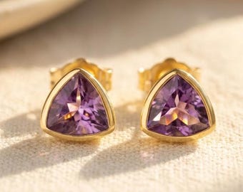 May include: A pair of gold stud earrings with triangular, purple amethyst stones. The earrings are set in a gold bezel setting, and the amethyst stones have a faceted cut. The earrings are displayed on a cream-colored surface.