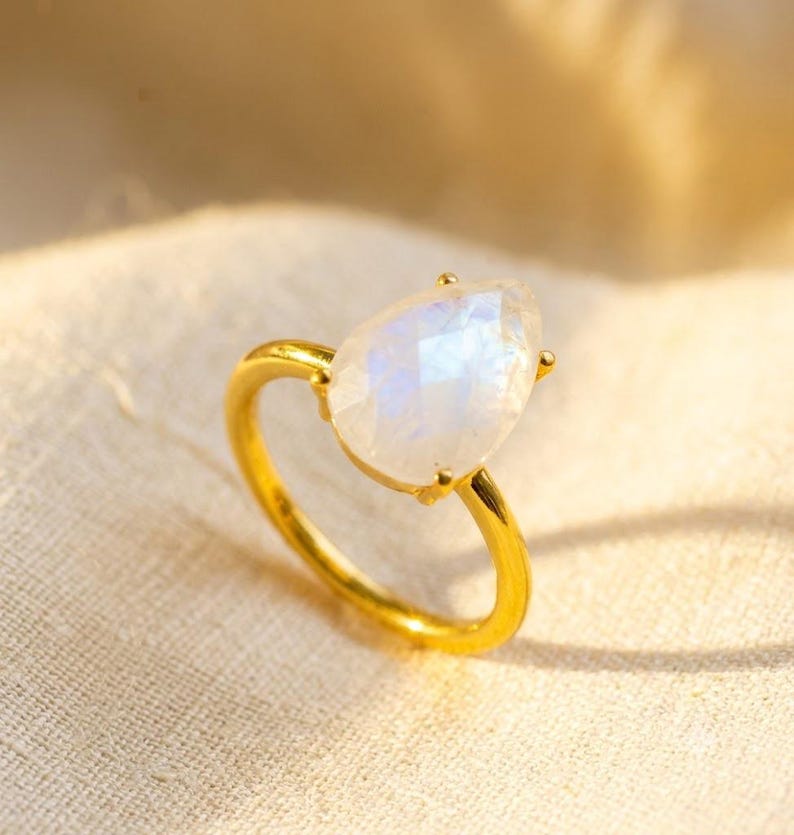 Gold moonstone teardrop ring displayed on soft fabric, showing the pear-shaped faceted rainbow moonstone and delicate prong setting. A June birthstone ring, gold moonstone ring, boho gemstone ring, birthday gift, anniversary gift, or special gift for her.