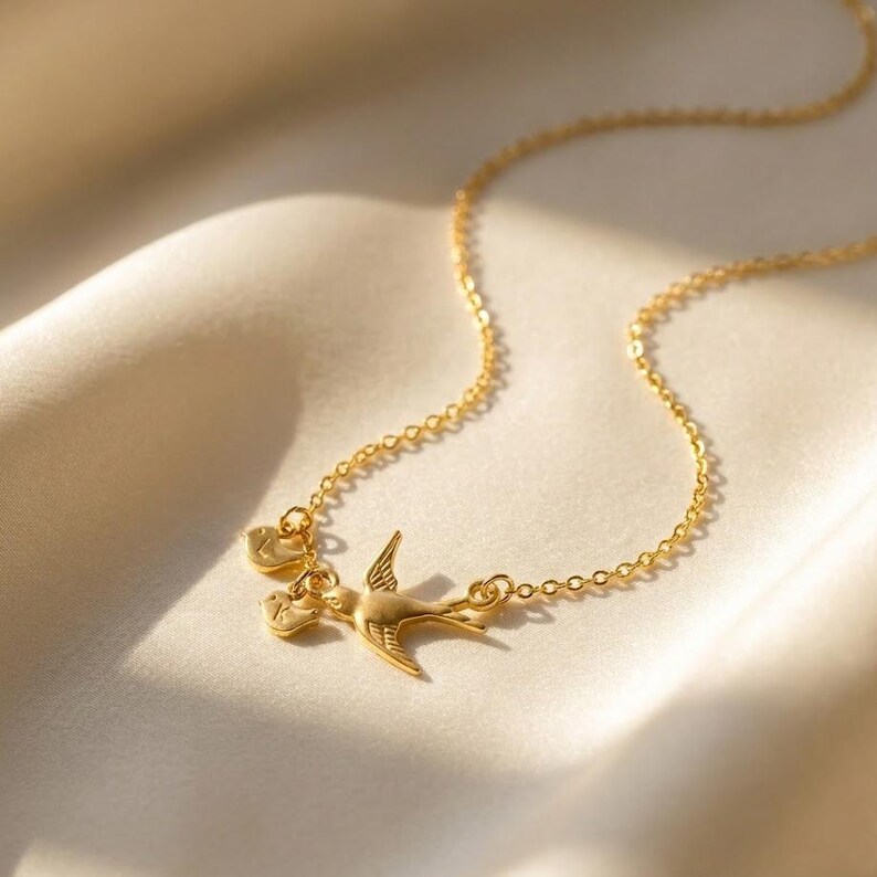 Gold mama bird necklace on linen with gentle shadows, close-up chain and charm arrangement, custom initial family necklace for mom or grandma.