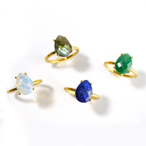 May include: Four gold rings with different gemstone settings. The rings feature a teardrop-shaped opal, a green gemstone, a blue gemstone, and a gray gemstone.