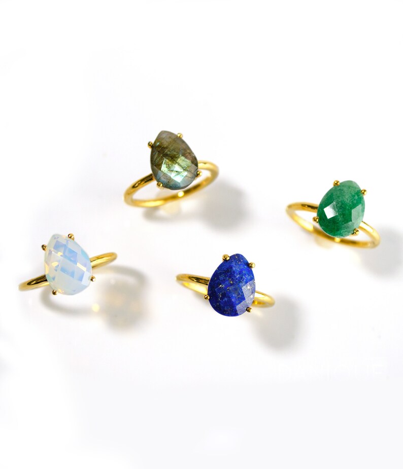 May include: Four gold rings with different gemstone settings. The rings feature a teardrop-shaped opal, a green gemstone, a blue gemstone, and a gray gemstone.