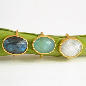 May include: Three gold rings with oval-shaped gemstones. The rings feature a blue labradorite, a green aventurine, and a white moonstone.
