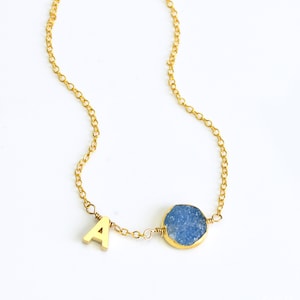 K&ouml;nnte beinhalten: Goldkette mit einem Anh&auml;nger aus blauem Achat und einem goldenen Buchstaben 'A'.