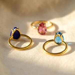 Moonstone ring and other teardrop gemstone rings shown on neutral fabric, featuring colorful pear-shaped stones in gold settings. A birthstone ring collection image for June moonstone rings, gemstone gifts, birthday jewelry, and matching birthstone pieces.